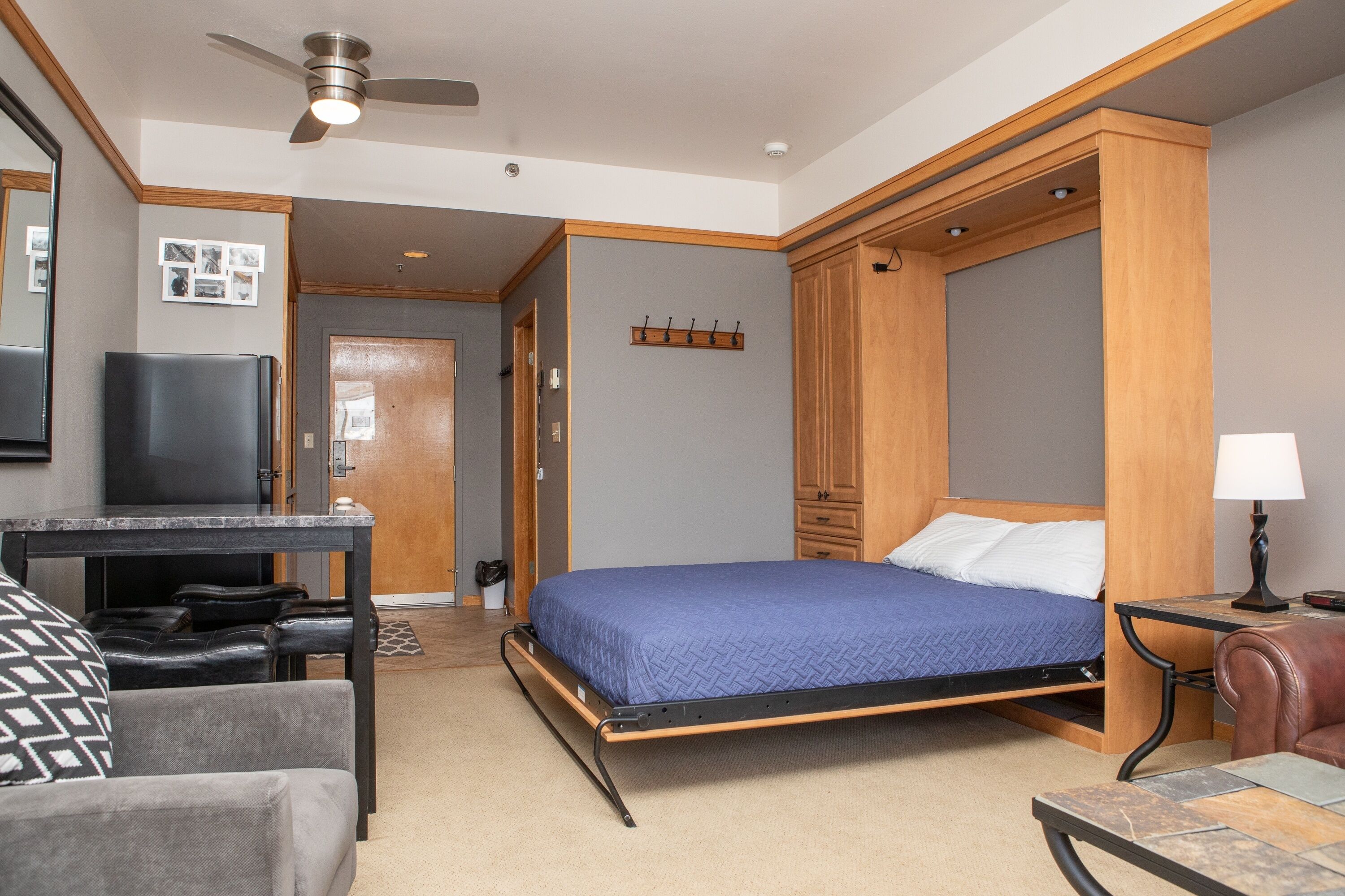 Une chambre d’hôtel de taille réduite, équipée d’un lit, d’un bureau, d’une chaise, d’une télévision et d’un ventilateur de plafond.
