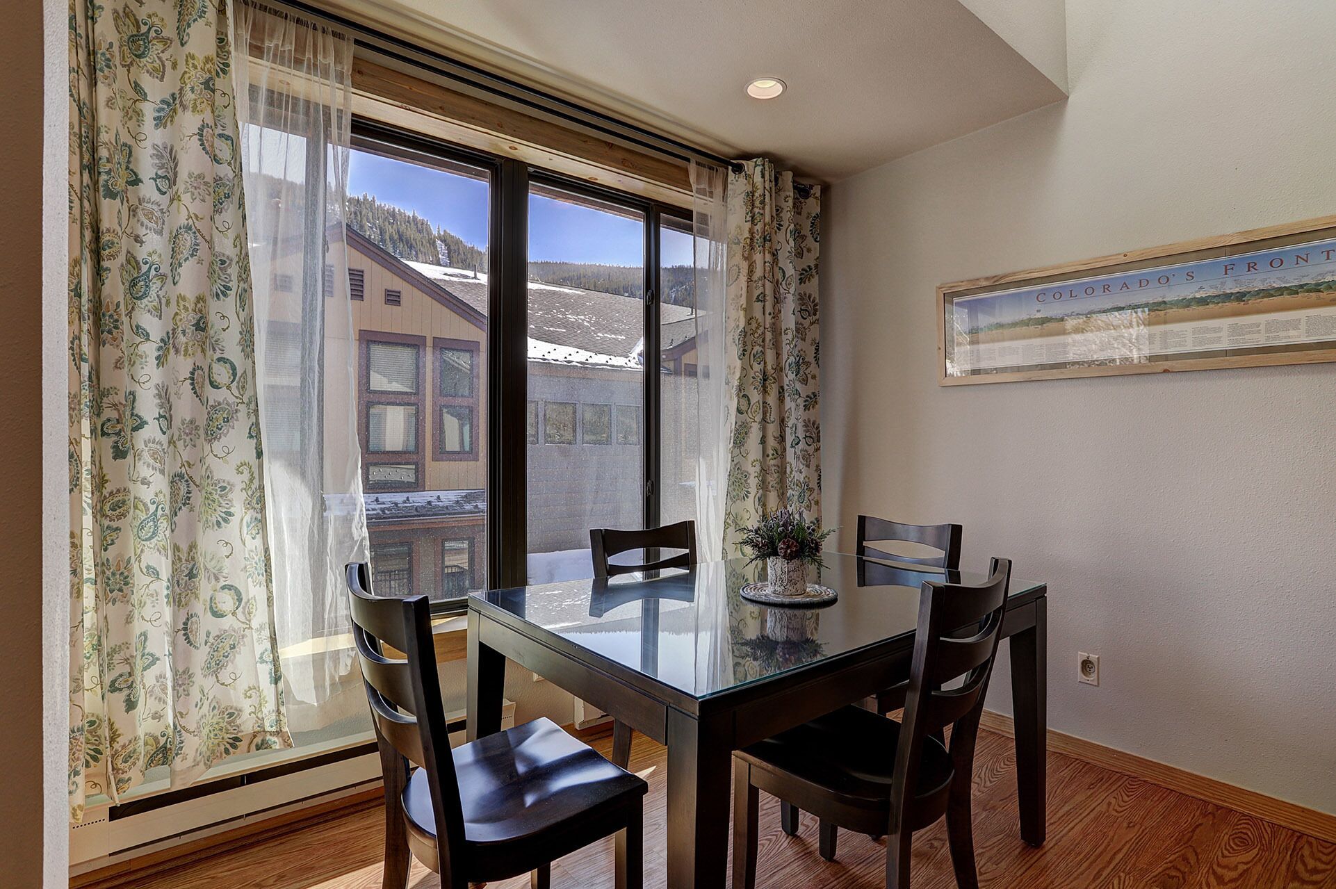 A dining area with a glass table and four chairs, large windows with curtains, and a framed picture on the wall.
