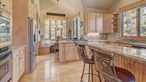 A spacious kitchen with wooden cabinets, a large island with bar stools, and a view of the outdoors through large windows. - 170 Elk Circle by RedAwning (Keystone)
