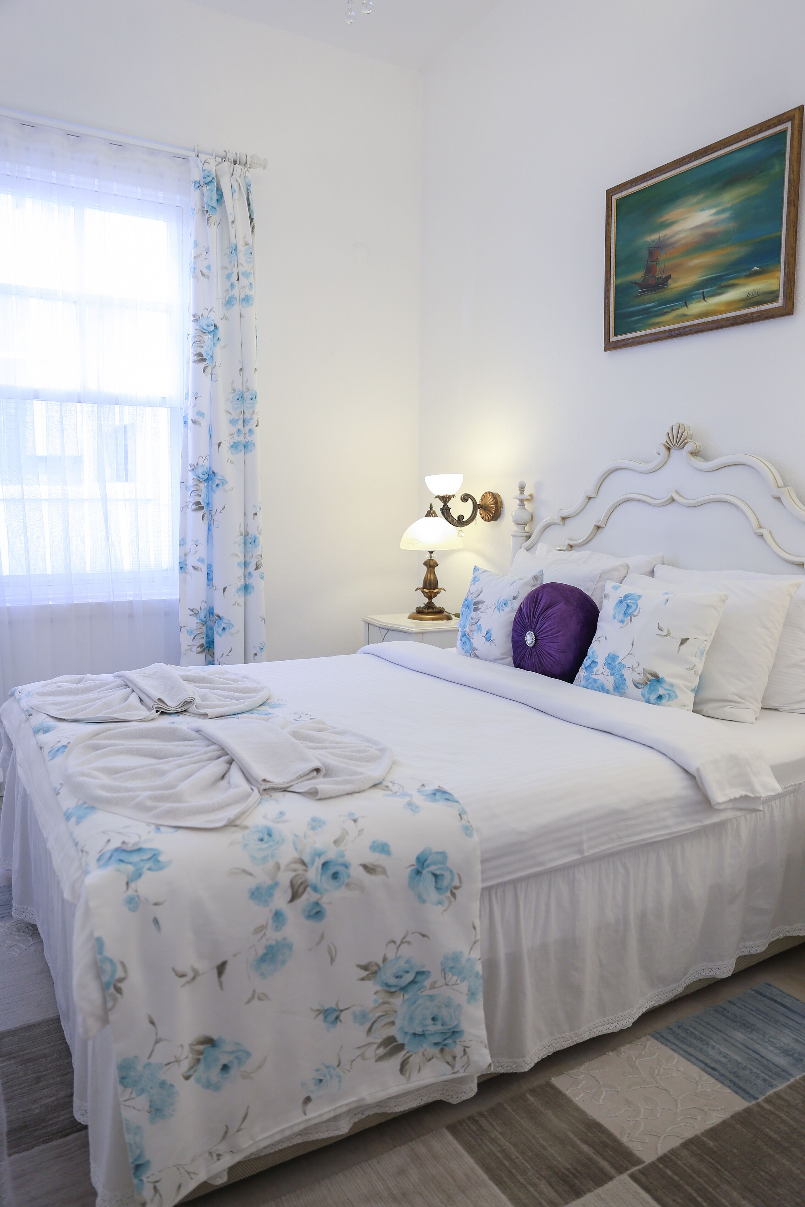 Un letto rifatto con lenzuola a motivi floreali, una lampada da comodino e un quadro appeso al muro.