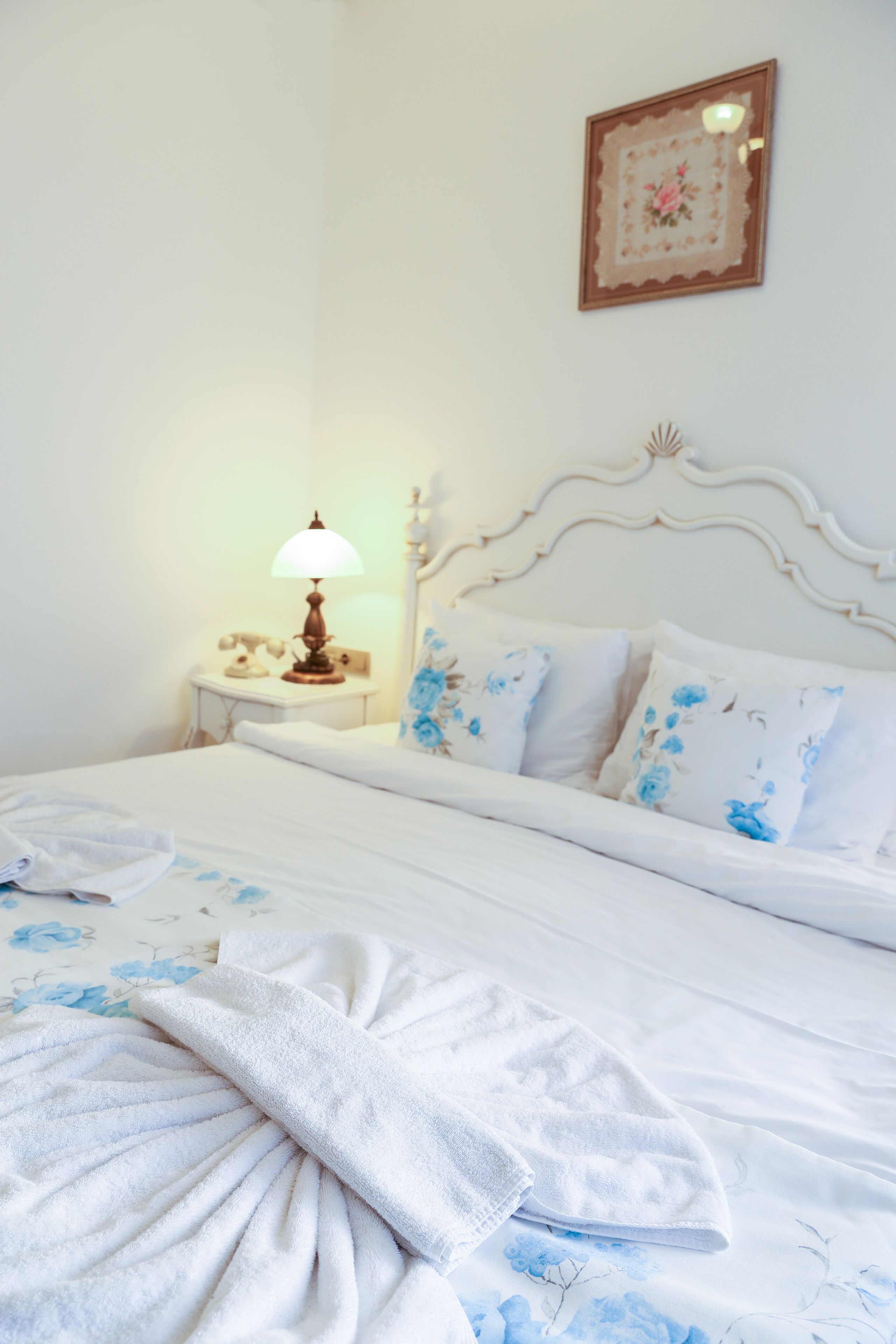 Una cama bien hecha con ropa de cama floral, una lámpara de noche y un cuadro en la pared.