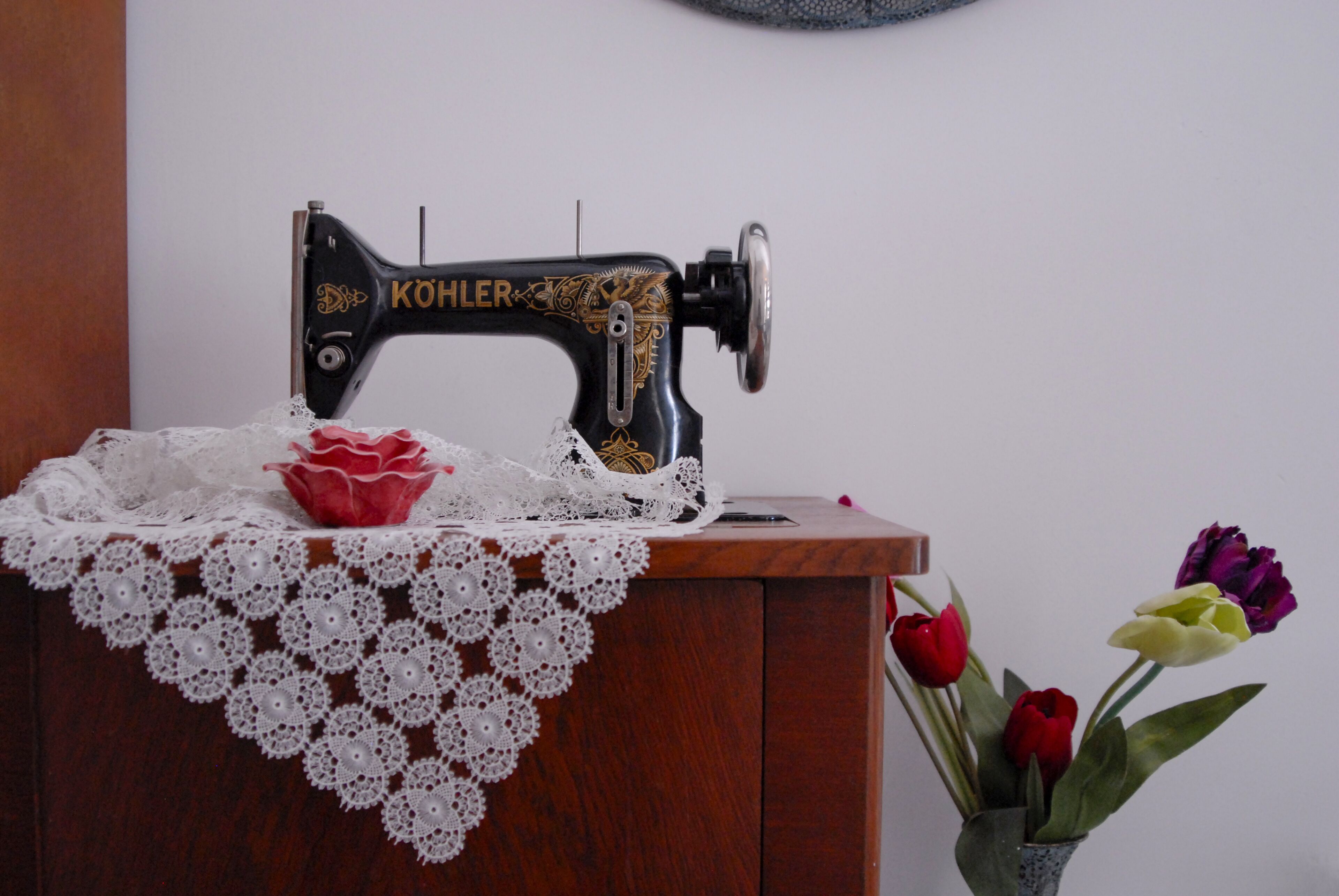 Una máquina de coser antigua sobre una mesa de madera con un mantel de encaje y un jarrón de tulipanes.