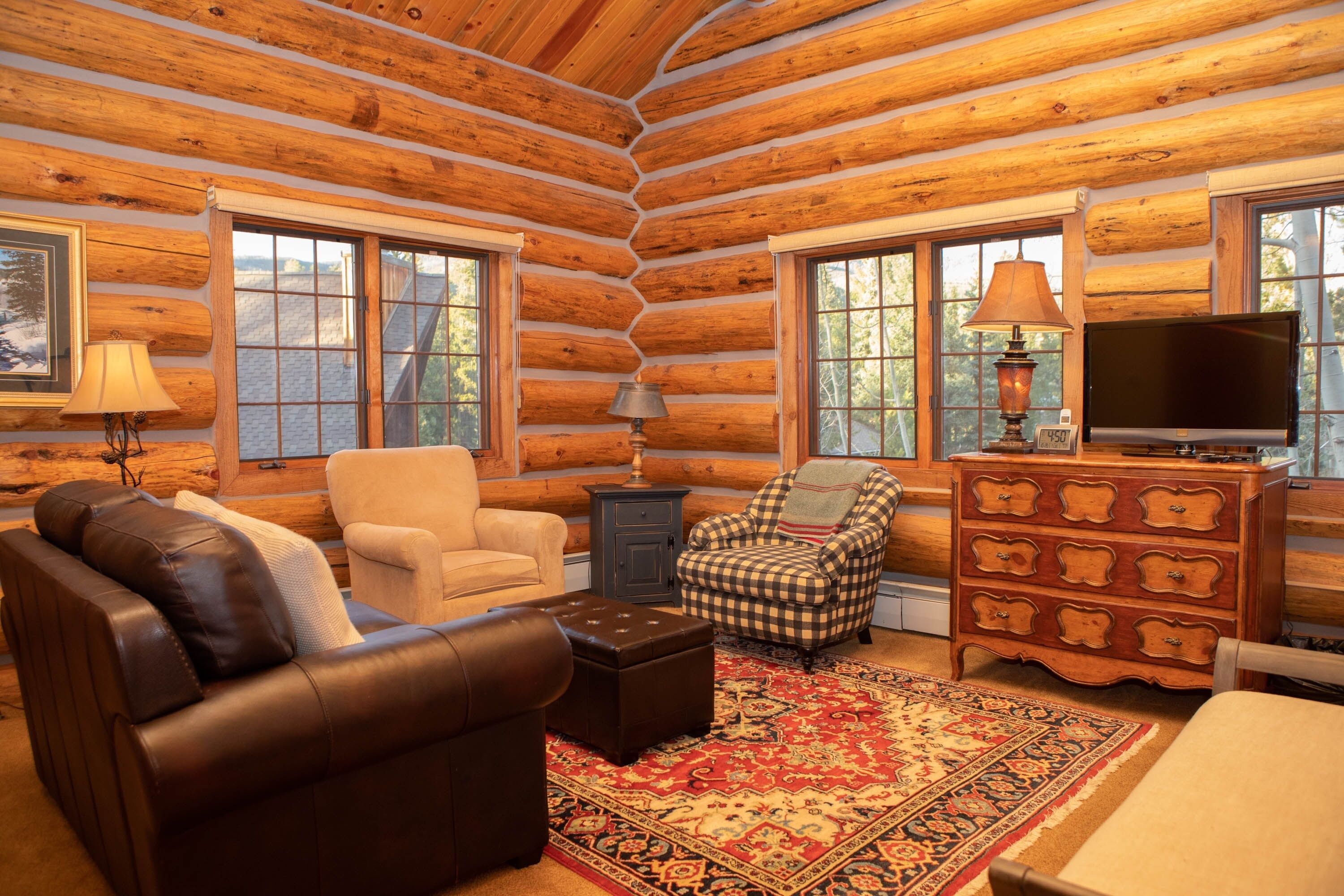 A log cabin interior with a brown leather sofa, a beige armchair, a plaid armchair, a wooden dresser, and a television.