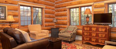 A log cabin interior with a brown leather sofa, a beige armchair, a plaid armchair, a wooden dresser, and a television.