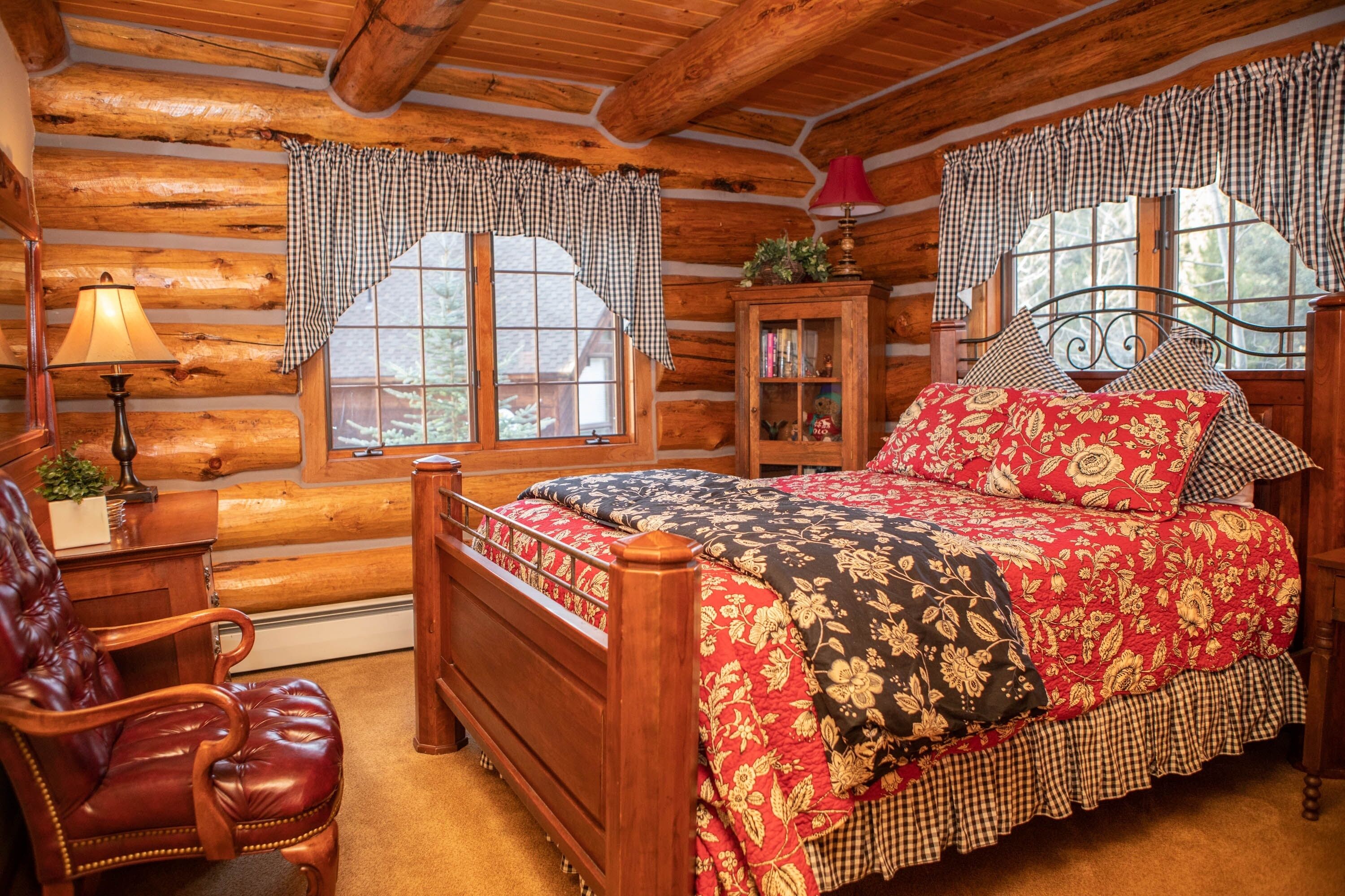 A log cabin bedroom with a wooden bed, a leather chair, a nightstand, and a window with checkered curtains.
