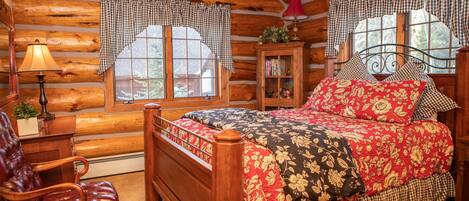 A log cabin bedroom with a wooden bed, a leather chair, a nightstand, and a window with checkered curtains.