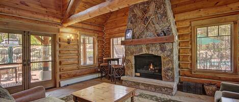 A log cabin interior with a stone fireplace, wooden furniture, and large windows.