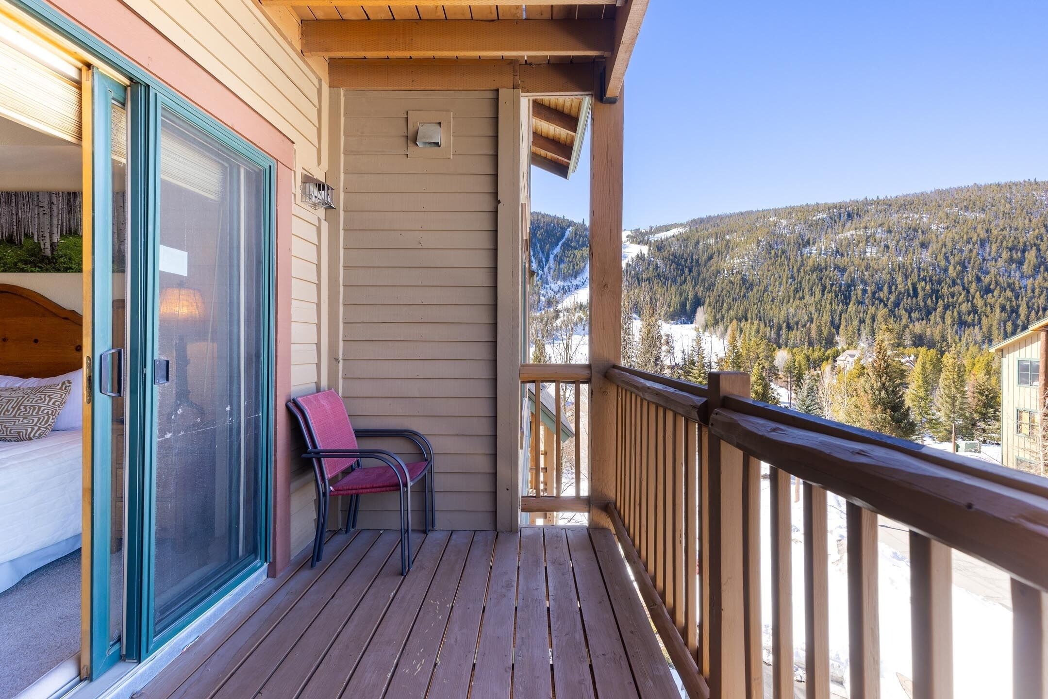 Un terrazza in legno con una porta vetrata che dà su una camera da letto, una sedia rossa e vista su un paesaggio montano innevato.