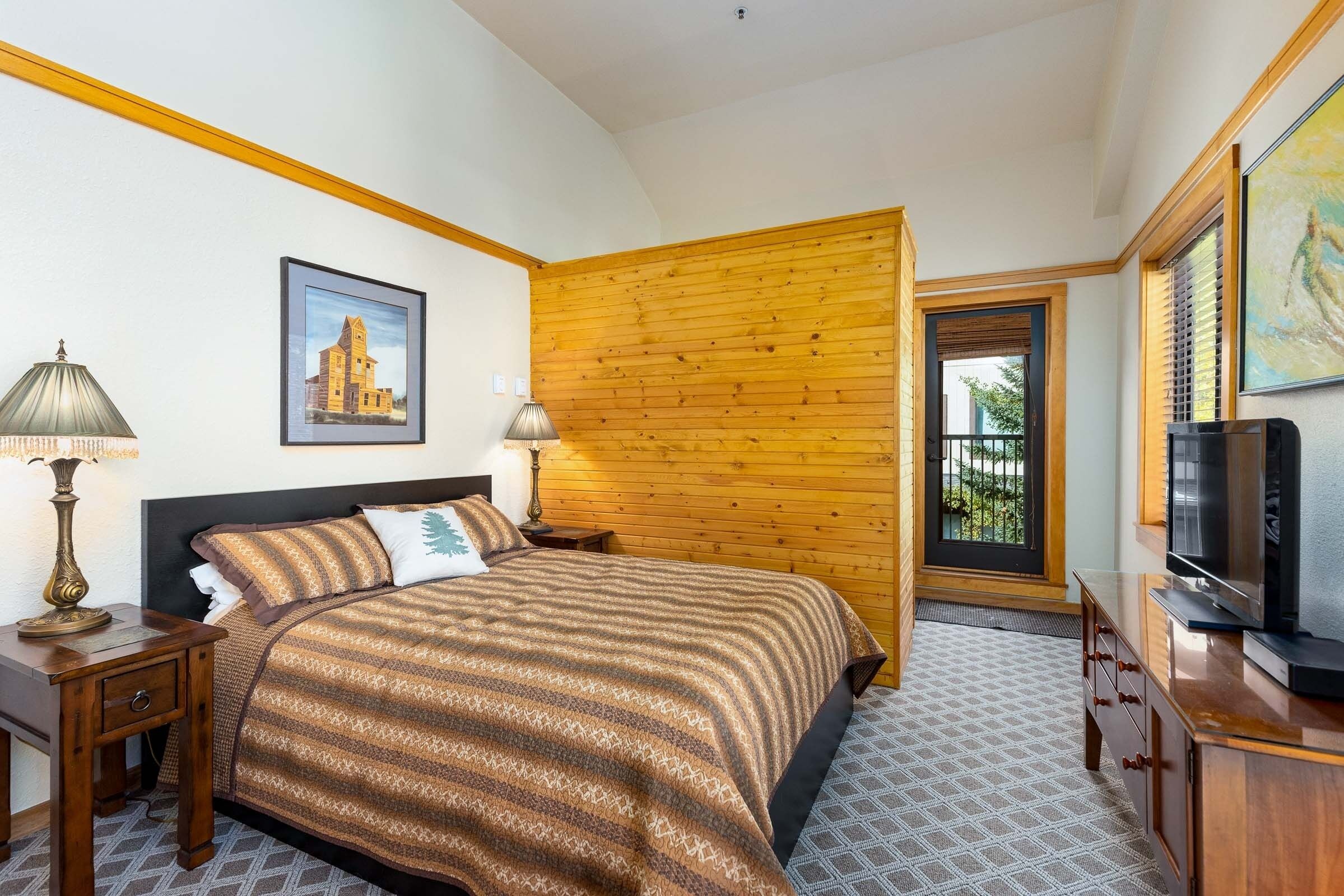 A bedroom with a bed, wooden panel wall, TV, and artwork.