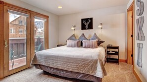 Une chambre avec un grand lit, une porte en bois, une vue sur les bâtiments à l’extérieur et un mur orné d’une œuvre représentant une tête de cerf.