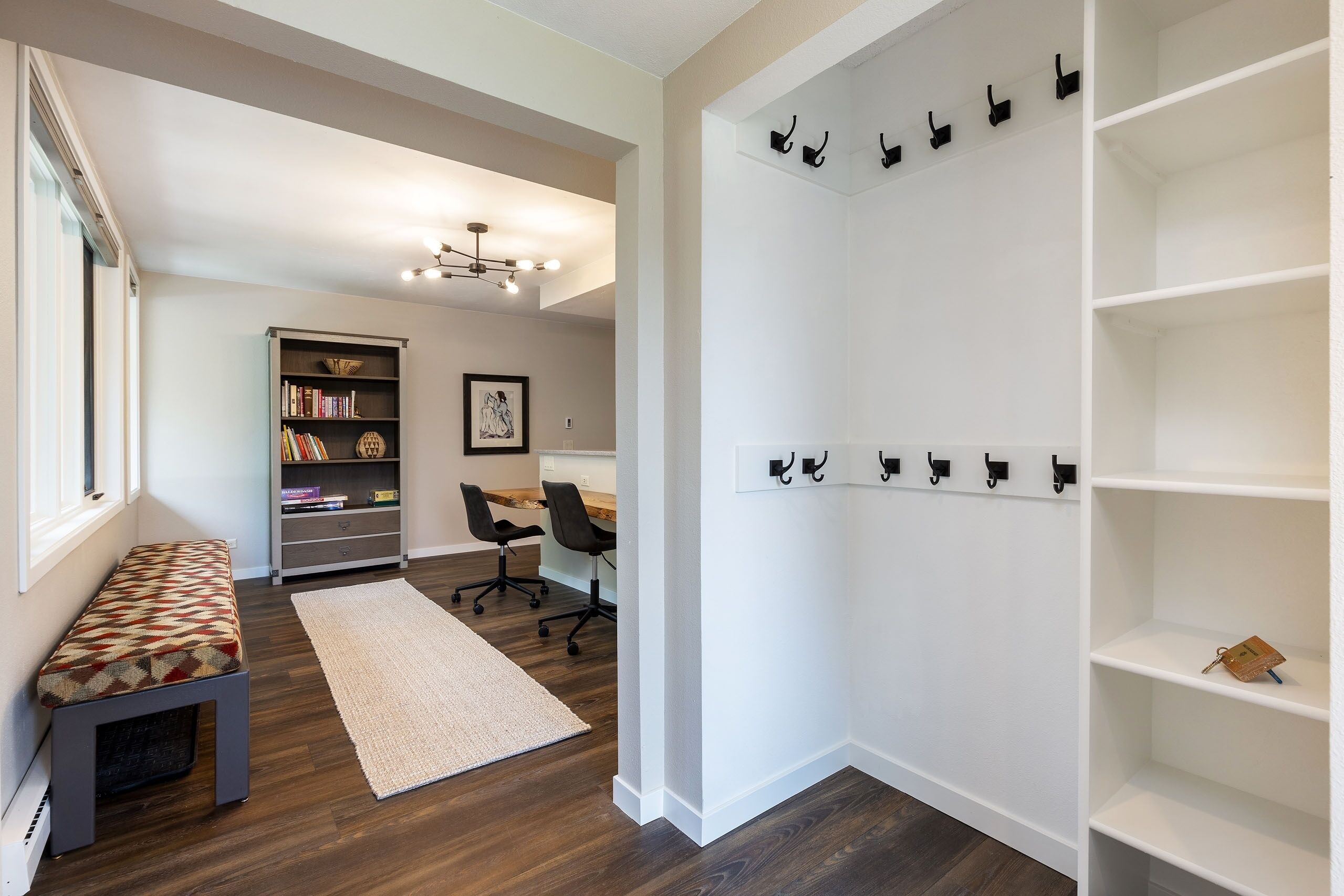 Una habitación moderna y bien iluminada, con piso de madera, una estantería, un escritorio con dos sillas, una pared blanca con perchero y un banco con diseño.