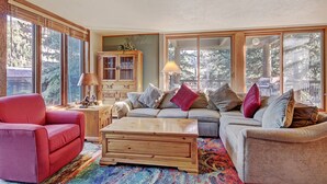 A cozy living room with a red armchair, a wooden coffee table, and a grey sofa with multiple cushions. There is a wooden cabinet with glass doors and a lamp on a side table. Large windows provide natural light and a view of trees outside. - 2019 Lodgepole 2 Bedroom Condo by RedAwning (Keystone)