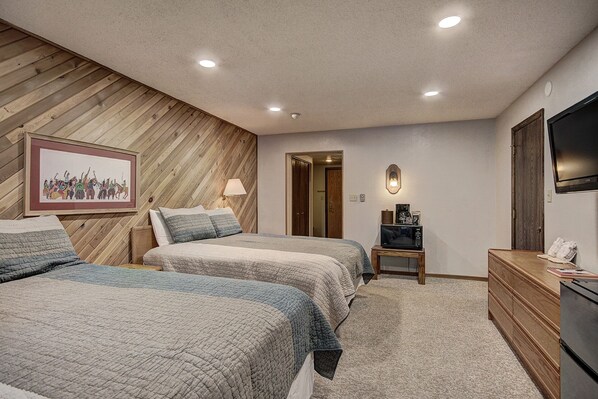 A bedroom with two beds, a wooden wall, a framed picture, a television, a microwave, and a dresser.