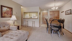 A living room with a sofa, a dining table with chairs, a kitchen area, and a hallway leading to a door.