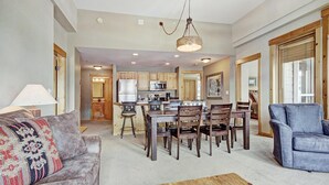 A spacious living room with a gray sofa, a blue armchair, and a wooden coffee table. A kitchen area with wooden cabinets and a dining table is visible in the background.