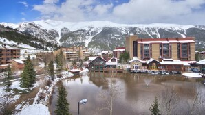 A snow-covered mountain town with a central building featuring large windows and a red roof, surrounded by smaller buildings and a body of water.