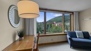 Une pièce avec une table à manger en bois, un canapé bleu, une grande fenêtre donnant sur des immeubles et des arbres, et un miroir fixé au mur.