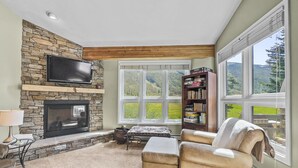 A cozy living room with a stone fireplace, a television mounted above it, a bookshelf, and a large window offering a view of the outdoors.