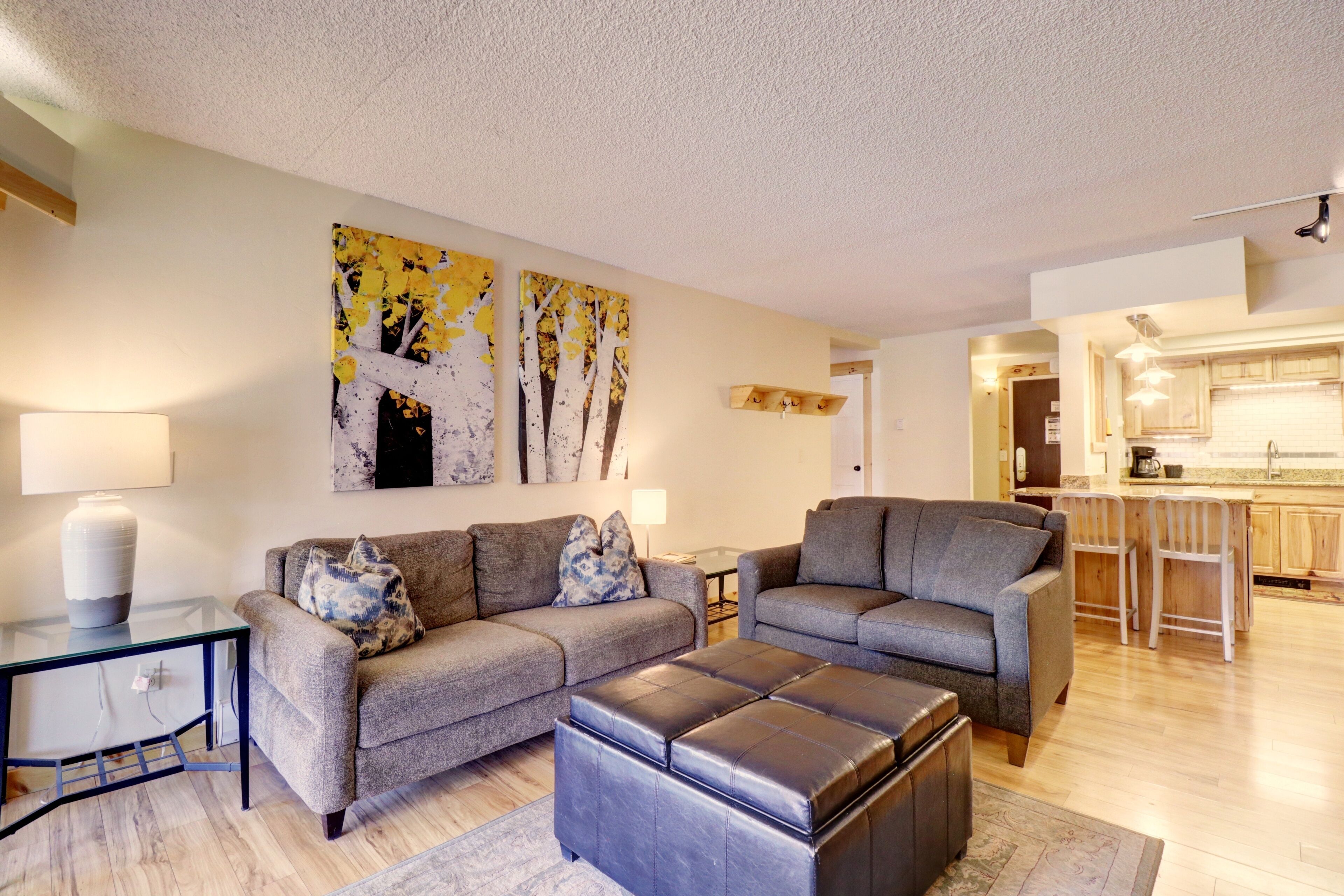 A modern living room with a sofa set, a glass coffee table, and a leather ottoman. There is a kitchen visible in the background.