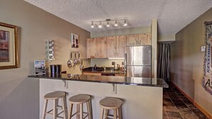 A kitchen with wooden cabinets, a stainless steel refrigerator, a granite countertop, and three wooden stools.