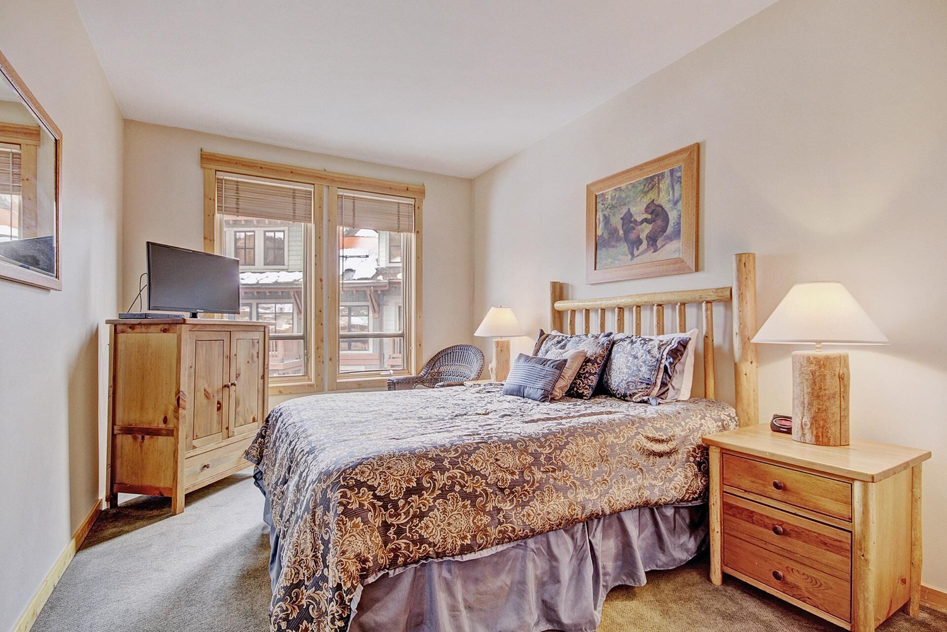 A bedroom with a bed, two bedside tables, a wooden cabinet, a television, and a window with blinds.