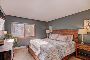 Quarto com cabeceira de madeira, colcha estampada, mesa de cabeceira com luminária e janela decorada com um floco de neve.
