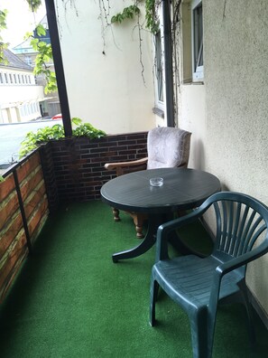 Une petite terrasse extérieure avec une table ronde et deux chaises, entourée d’un mur de briques et d’un mur blanc percé d’une fenêtre.