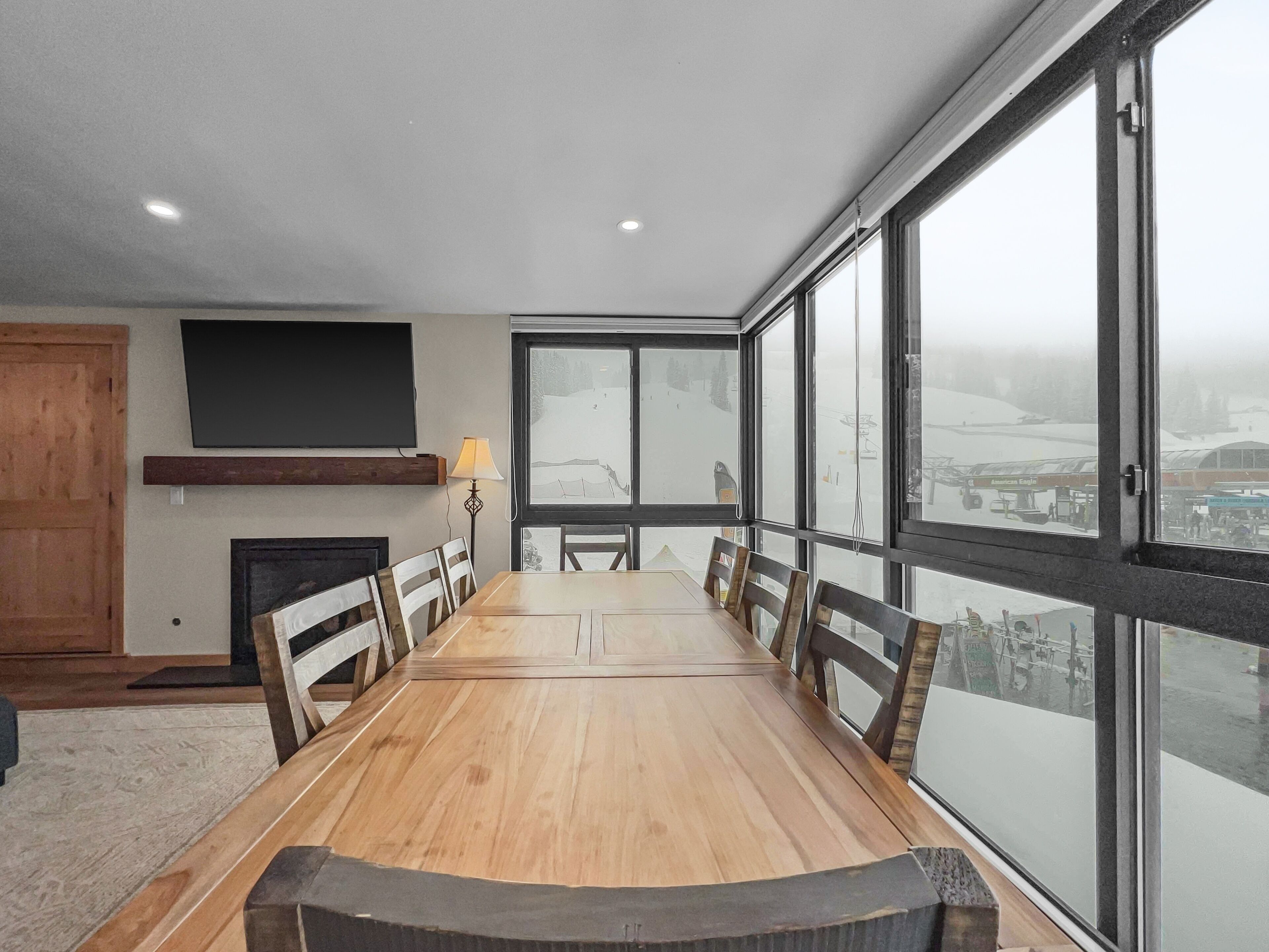 Uma sala de jantar com uma grande mesa de madeira, cadeiras, lareira, televisão fixada na parede e janelas amplas com vista para uma paisagem nevada.