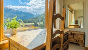 Ein Zimmer mit Schreibtisch aus Holz, einem Sessel, einer Kommode und einem großen Fenster mit Blick auf die Berge.