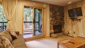 Ein gemütliches Wohnzimmer mit Kamin aus Stein, einem wandmontierten Flachbildfernseher, einer Couch mit gemusterten Kissen und einer Glastür zu einer Terrasse.