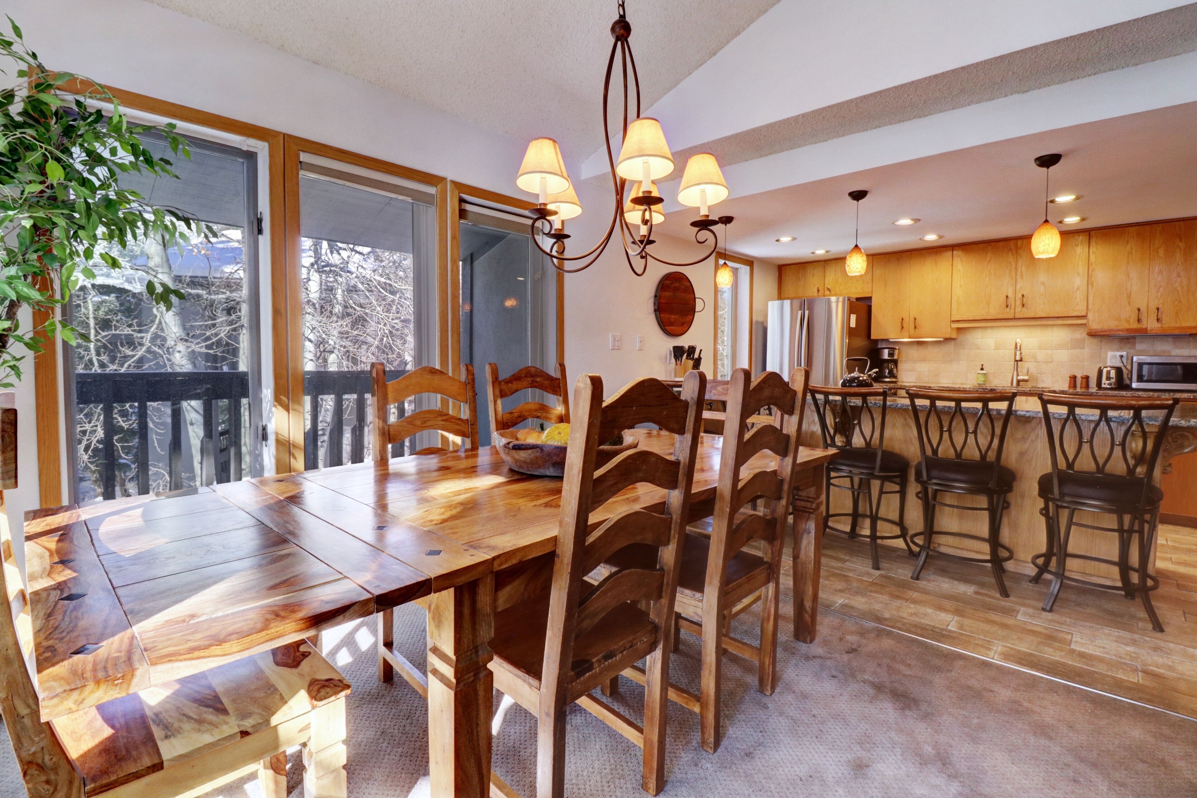 A dining area with a wooden table and chairs, a chandelier, and a view through large windows.