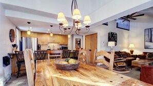 Ein geräumiges Wohnzimmer mit einem großen Esstisch aus Holz, einer Küche im Hintergrund und einem Deckenventilator.