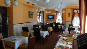 Un restaurante con varias mesas y sillas, un televisor montado en la pared y un ventanal con cortinas.