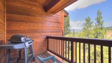 A wooden deck with a grill and chairs, overlooking a forested landscape.