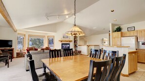 Amplia sala de estar con un gran comedor de madera, asientos cómodos, televisor y una cocina con armarios de madera y refrigerador.