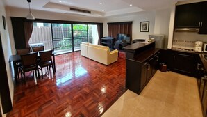 A modern living room with a sofa, dining table, and chairs, a kitchen with a stove and microwave, and a large window with a view of greenery.