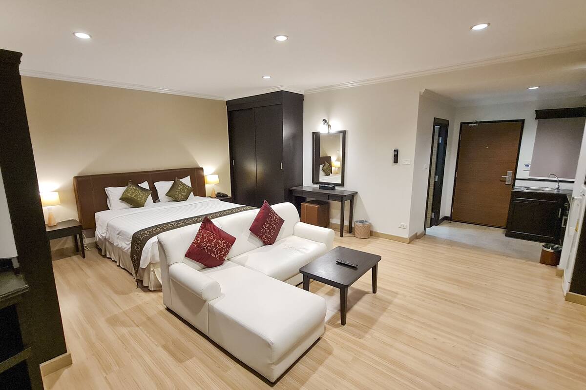 Une chambre d’hôtel comprenant un lit, un canapé, une petite table, un bureau, un miroir, une armoire et une kitchenette.