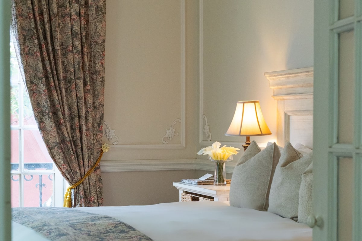 A neatly made bed with a floral bedspread, a bedside lamp, and a nightstand with a vase of flowers.