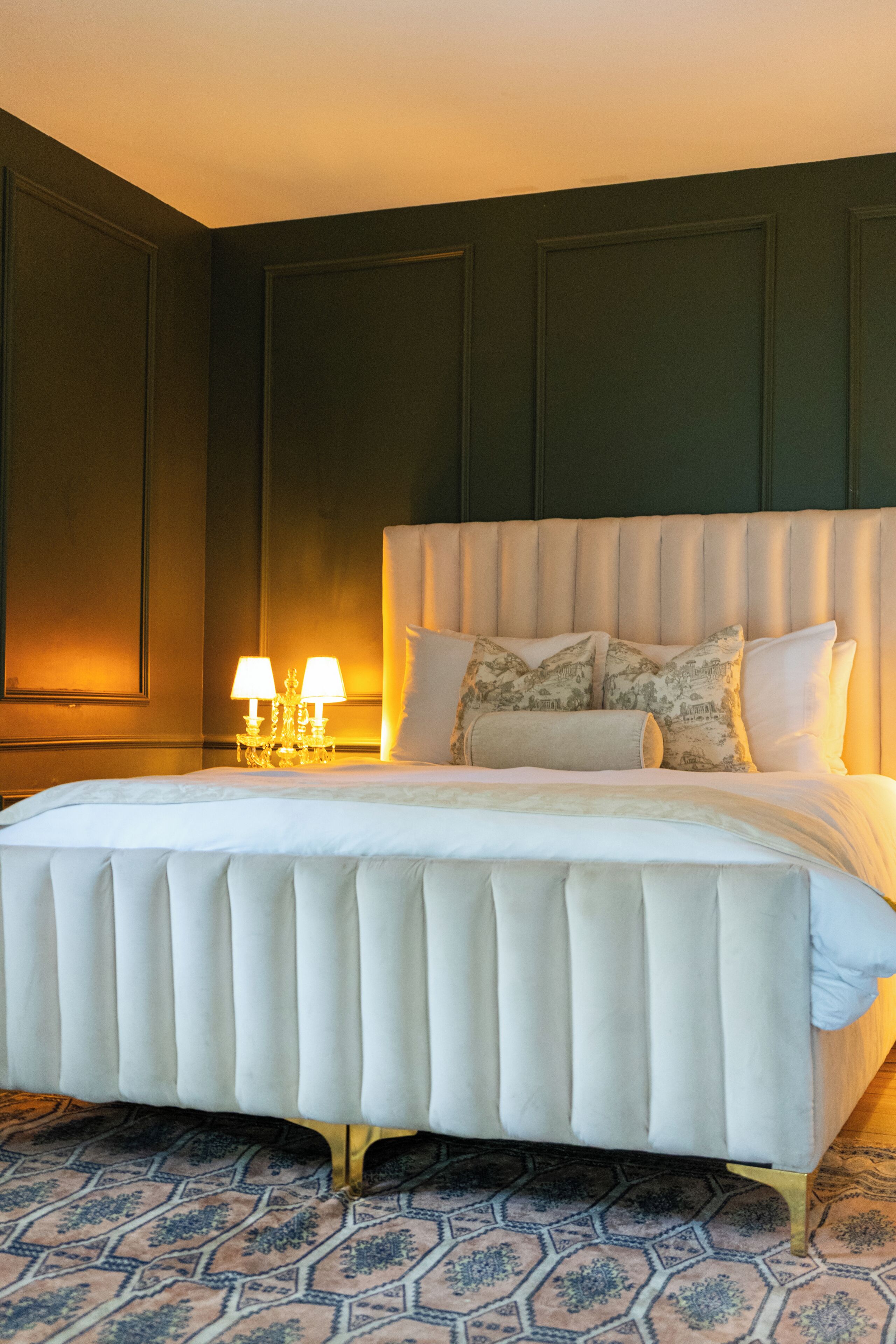 A large bed with a tufted headboard, a bedside table with two lamps, and a patterned rug.
