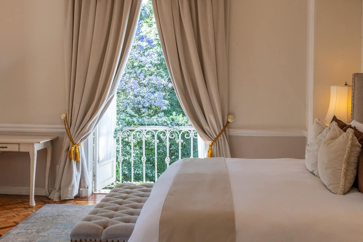A bedroom with a large bed, a chandelier, curtains, and a view of greenery outside.