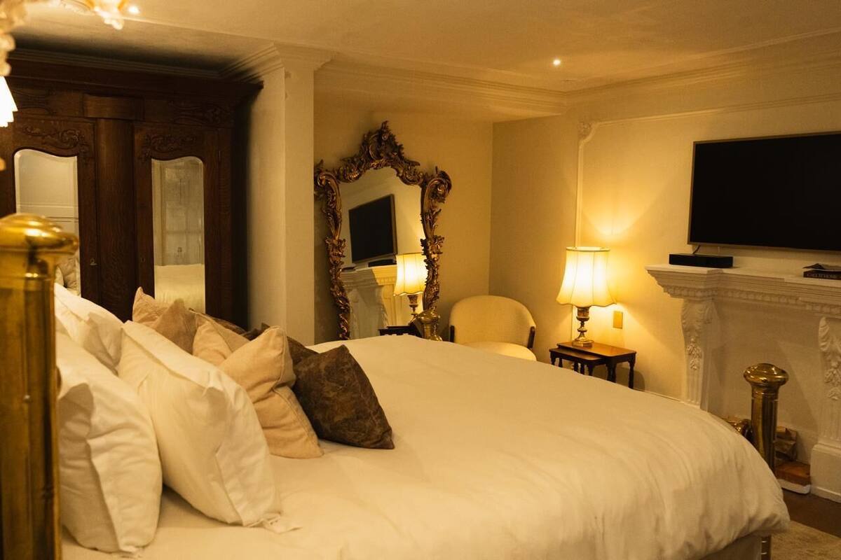 A hotel room with a large bed, a mirror, a television, and a fireplace.
