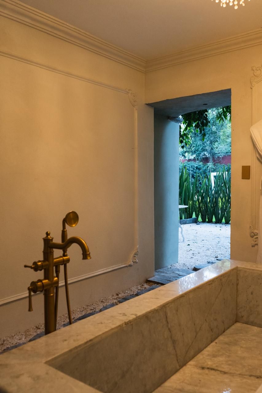 A bathroom with a marble tub, a gold faucet, and a view of a garden through an archway.