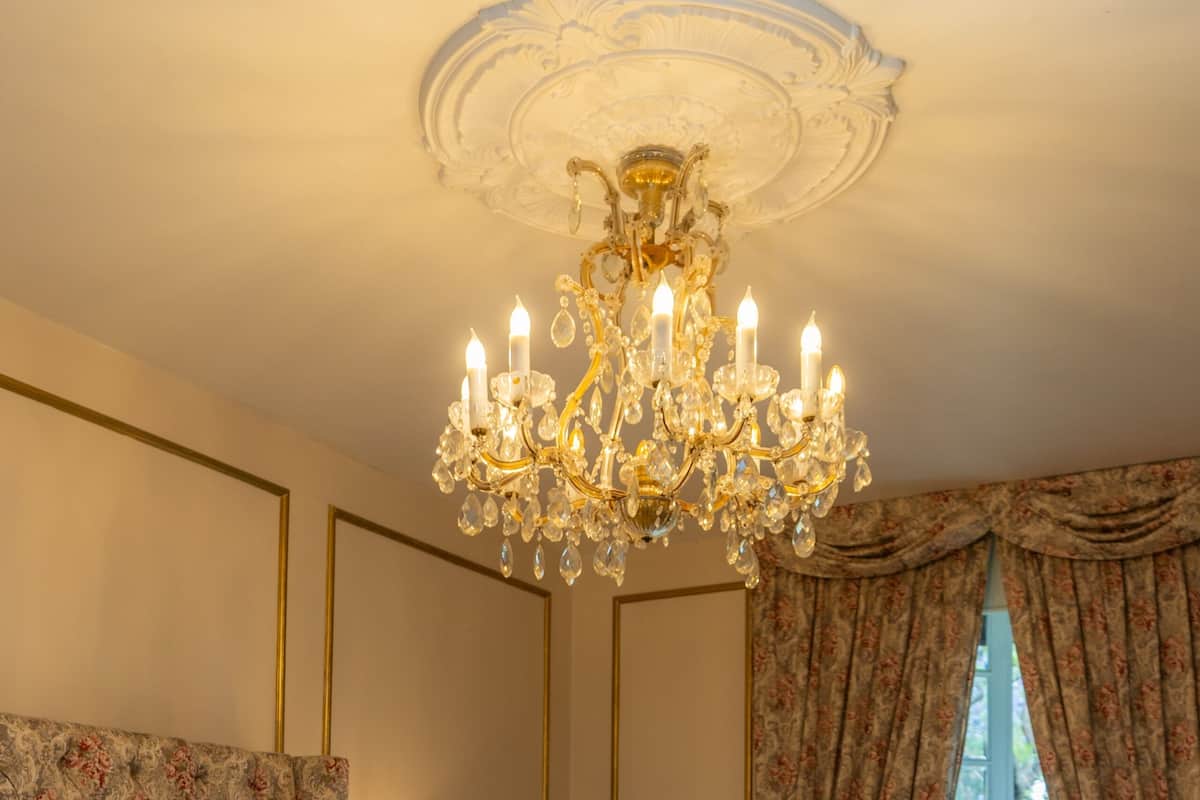 A bedroom with a large bed, a chandelier, floral-patterned headboard, a nightstand, and a view of greenery outside.