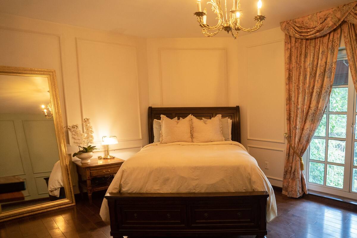 A bedroom with a large bed, a nightstand with a lamp, a mirror, and a window with curtains.