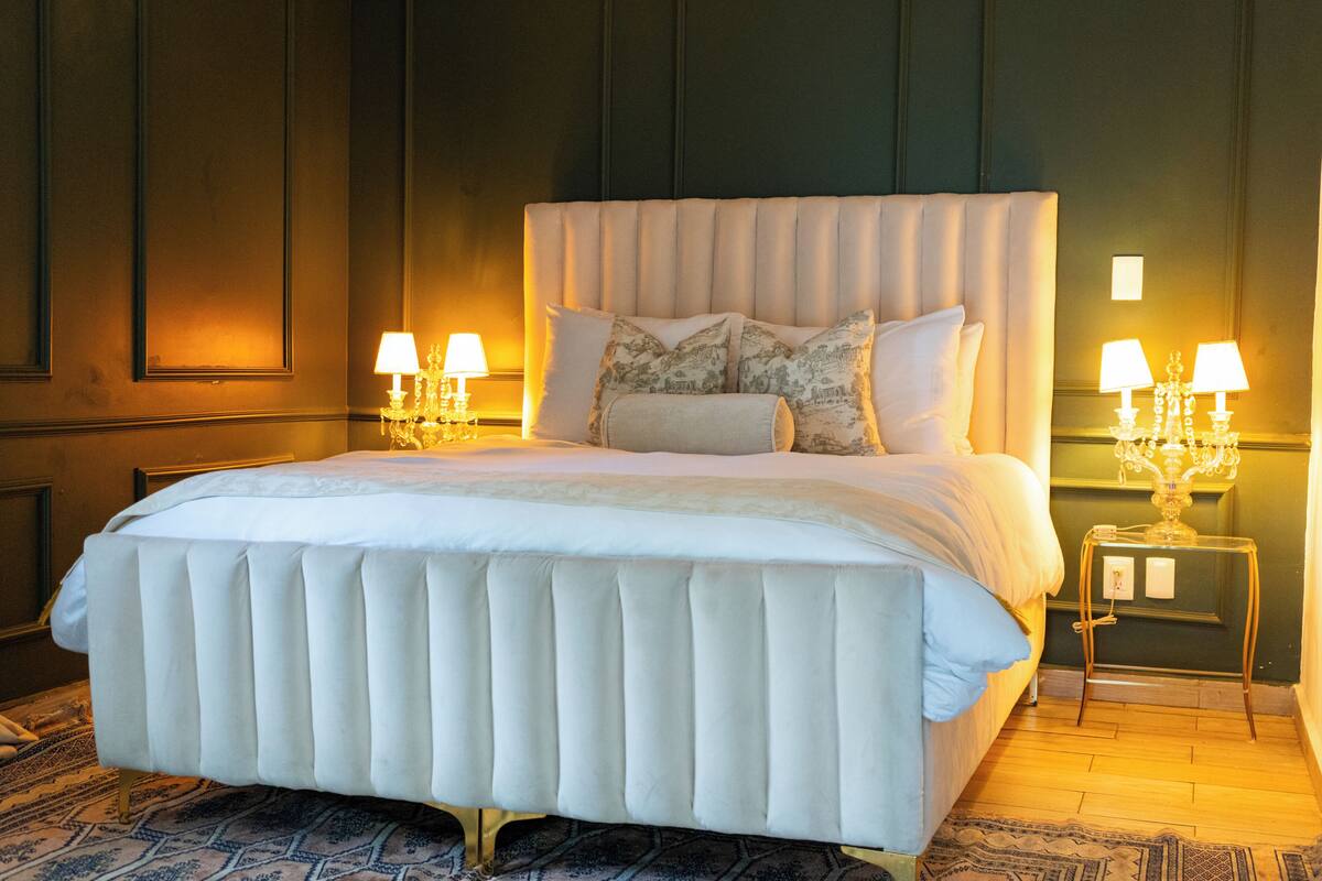 A large bed with a tufted headboard, two bedside tables with lamps, and a patterned rug on the floor.