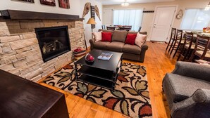 A cozy living room with a stone fireplace, a sofa with red cushions, a coffee table, and a dining area with wooden chairs.