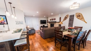 A modern kitchen with a built-in dishwasher, a dining area with wooden chairs, and a living room with a sofa and fireplace.