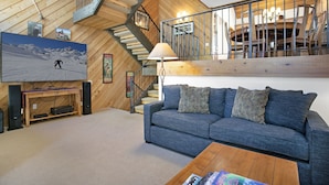 A living room with a blue sofa, wooden coffee table, and a TV displaying a ski scene. A staircase with a metal railing leads to an upper level with a dining area. - 188 Ridgepoint 3 Bedroom Townhouse by RedAwning (Avon)