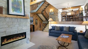 A spacious living room with a stone fireplace, a blue sofa, and a wooden staircase leading to an upper level. - 188 Ridgepoint 3 Bedroom Townhouse by RedAwning (Avon)