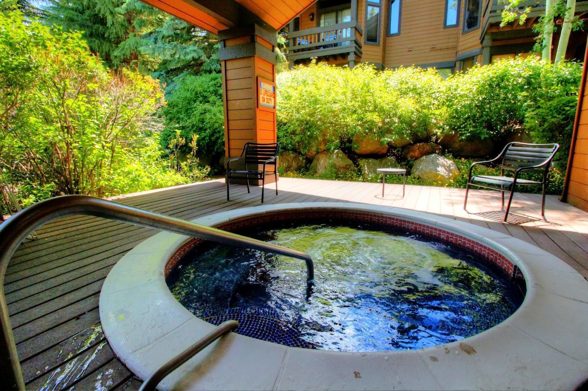 A hot tub with a wooden deck, surrounded by greenery and a building with balconies.